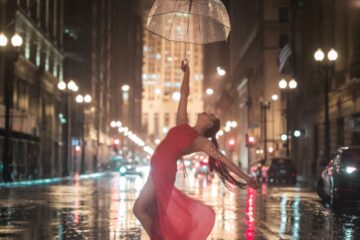 A Day in the Rain… Never the Same Nighttime urban scene with a dancer in a red dress posing gracefully on a wet street.